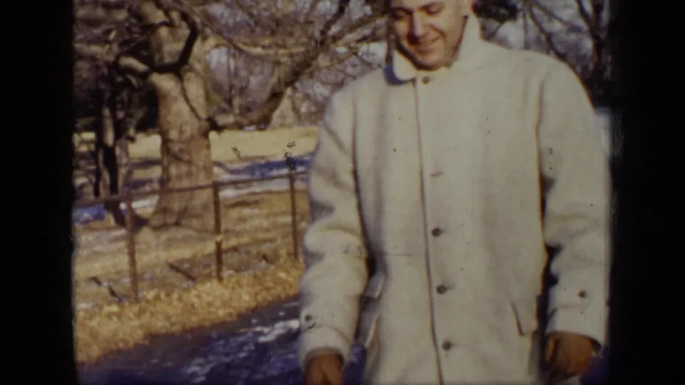 A close frame shows the man in a buttoned light winter coat, with the park path, fence, bare trees, and snow patches in the background.