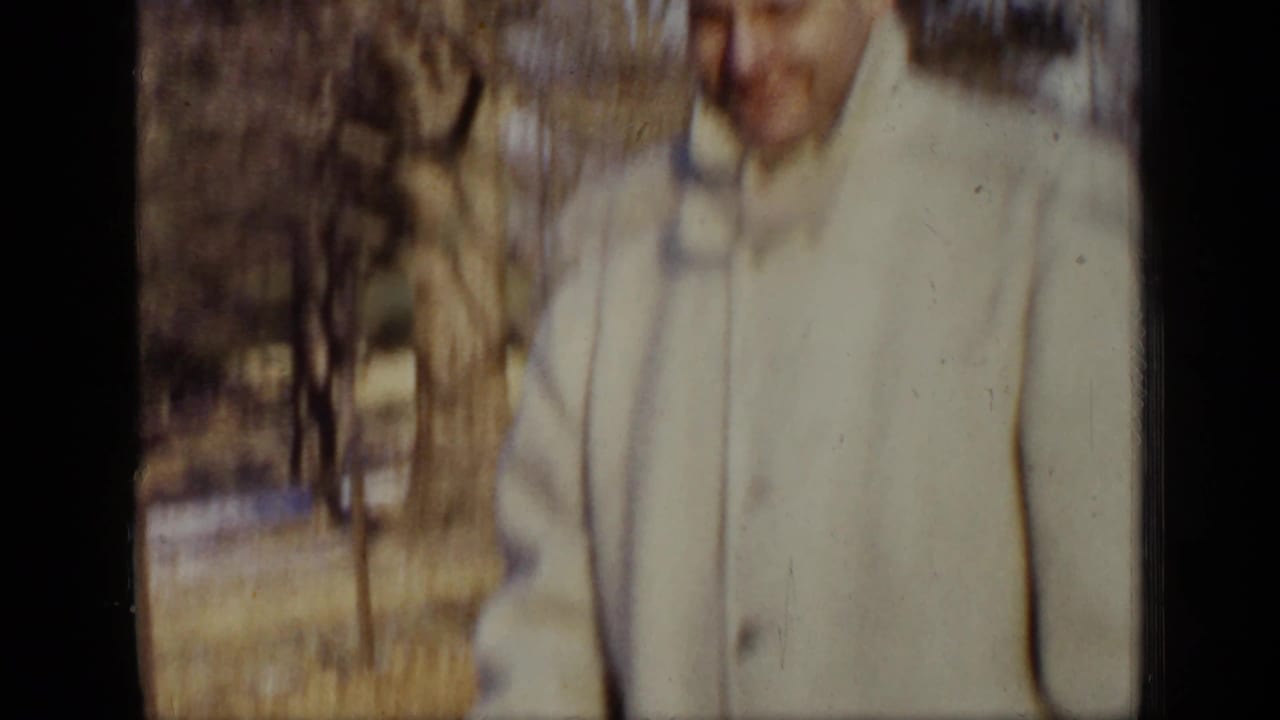 Adult man in a light coat standing outdoors near bare trees.