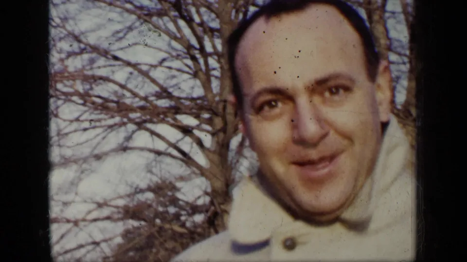 Close view of a smiling adult man in a light coat, filmed outdoors against bare winter trees in New York.