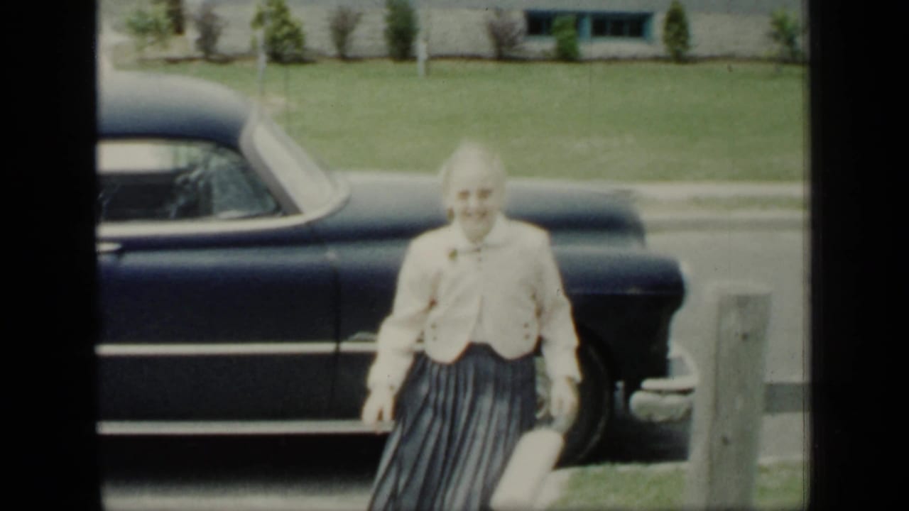 Woman in a light jacket and pleated skirt walking past a dark sedan on a residential street.