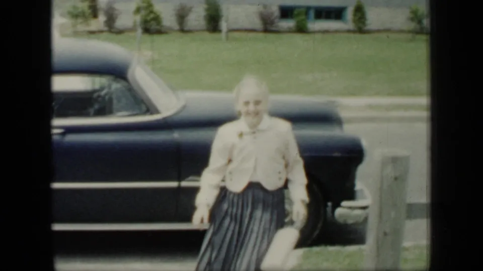 In 1948 New York home-movie footage, a woman in a light jacket and pleated skirt approaches the camera beside a dark 1940s sedan.