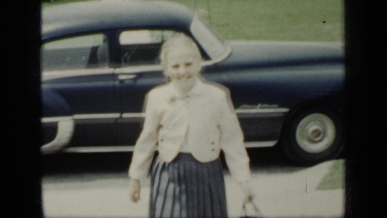 Smiling woman in a light jacket standing near a dark sedan with houses and lawn behind her.