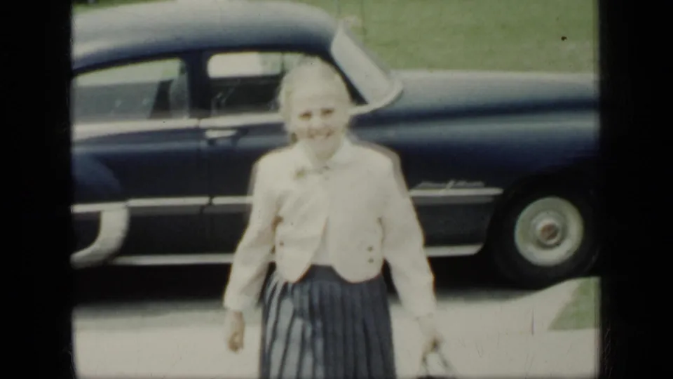A smiling woman stands near a dark sedan on a New York residential street, with lawns and houses visible behind her.