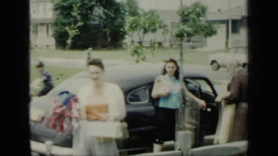 Women in period dress carry parcels or gifts near a dark sedan on a suburban New York street in 1948 home-movie footage.