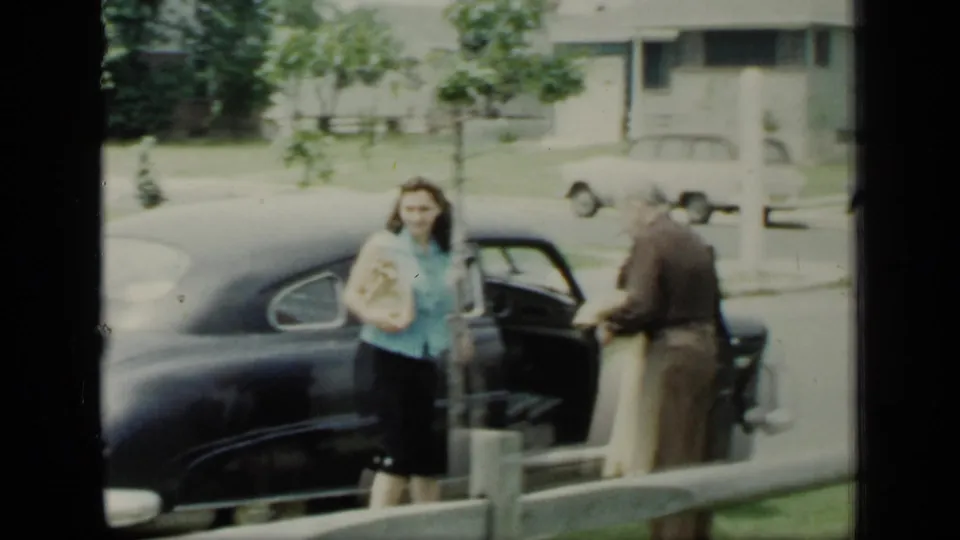 A woman in a blue blouse holds a parcel near an open car door while a man stands by the sedan on a suburban New York street.