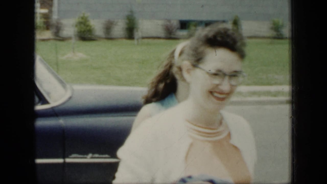 In 1948 New York home-movie footage, a smiling woman with glasses and a light cardigan stands near a dark car on a suburban street.