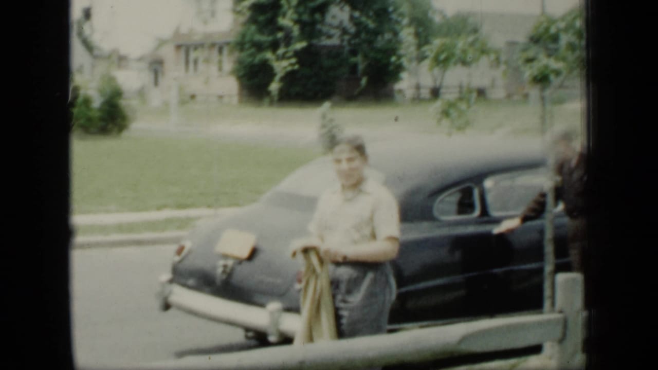 Adult carrying a coat stands by a dark car on a residential street.