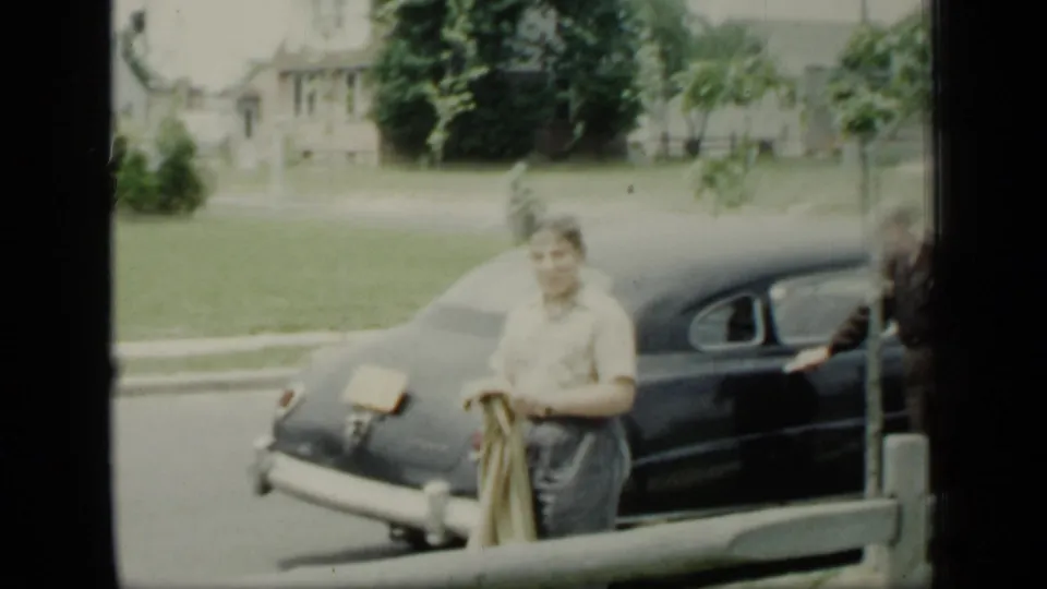 An adult carrying a coat smiles near a dark rounded car on a New York suburban street in 1948 home-movie footage.