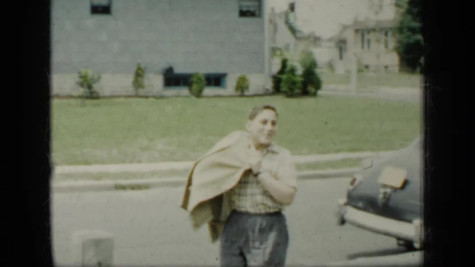 The adult smiles toward the camera while holding a coat near a dark vintage car, with lawn and buildings behind.