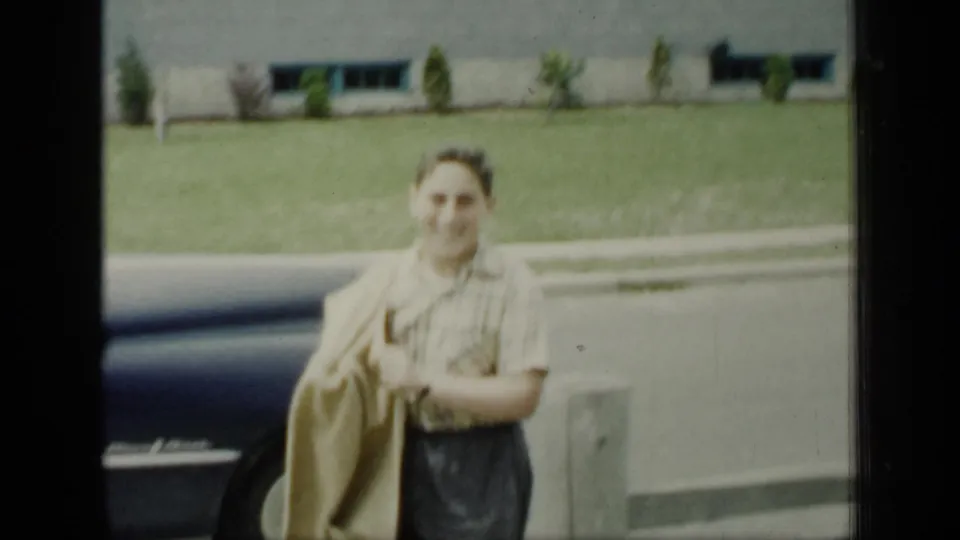 A smiling adult holding a coat stands in front of a dark car, with grass and a low building visible in the background.
