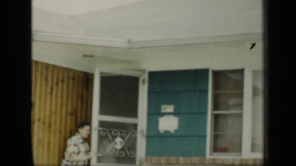 1948 New York exterior frame with a woman moving along the porch near a teal door, windows, and light wood siding.