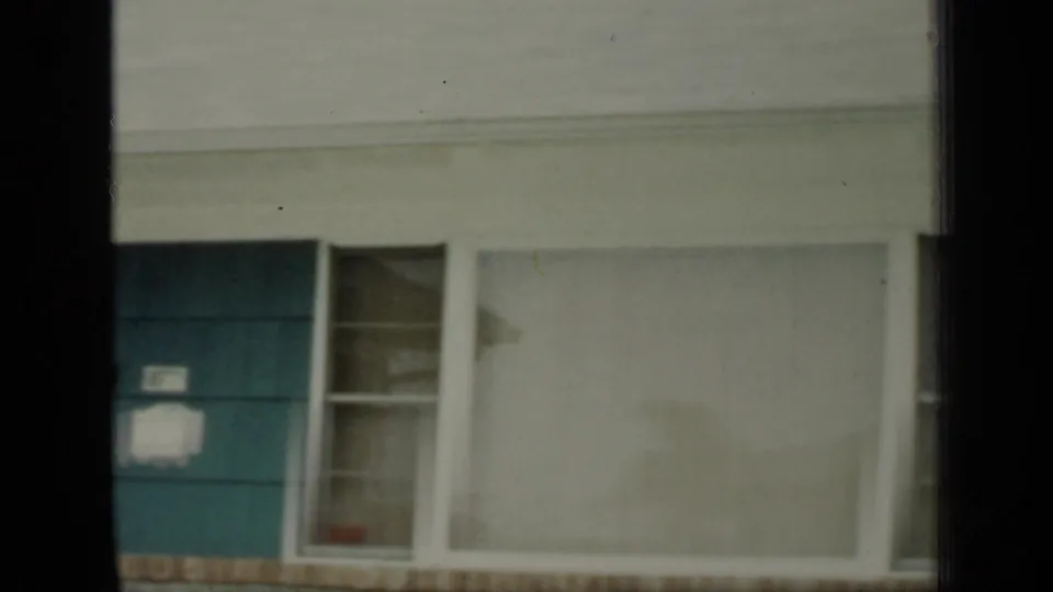 1948 New York home exterior with a teal door, porch window, broad front window, pale siding, and brick foundation trim.