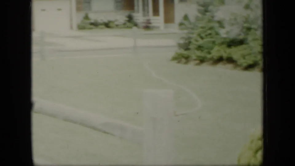 Residential front yard in 1948 New York with lawn, shrubs, a small white fence, and house exterior visible in faded 8mm color.