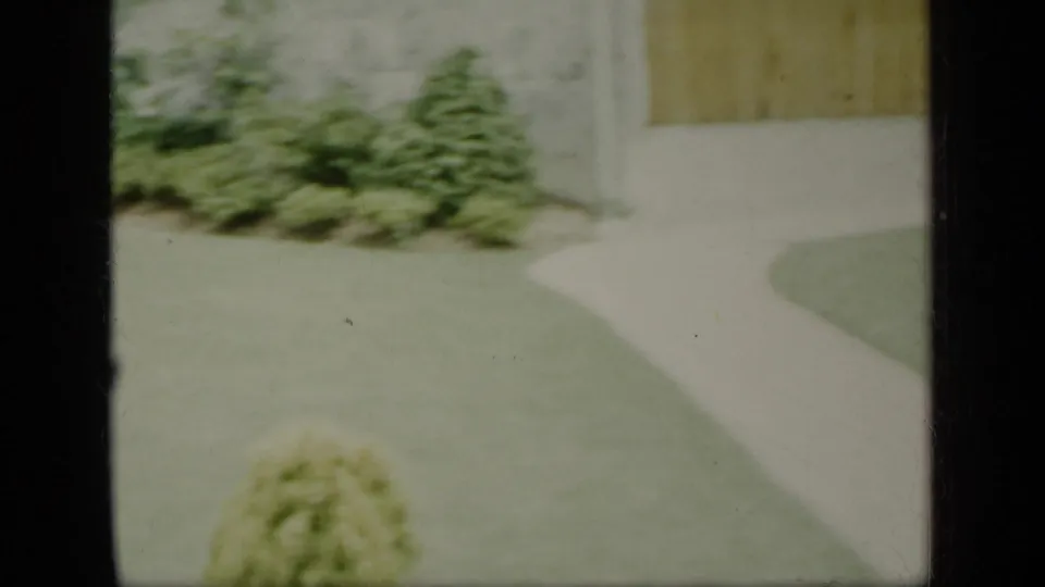 Soft 8mm view of a New York residential lawn, curved walkway, shrubs, and garage-side exterior, dated by metadata to 1948.