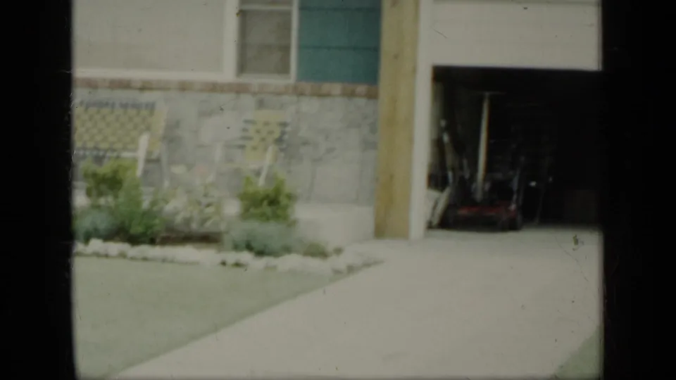 1948 New York 8mm frame with porch chairs, house siding, stone foundation, lawn, and small garden plants.