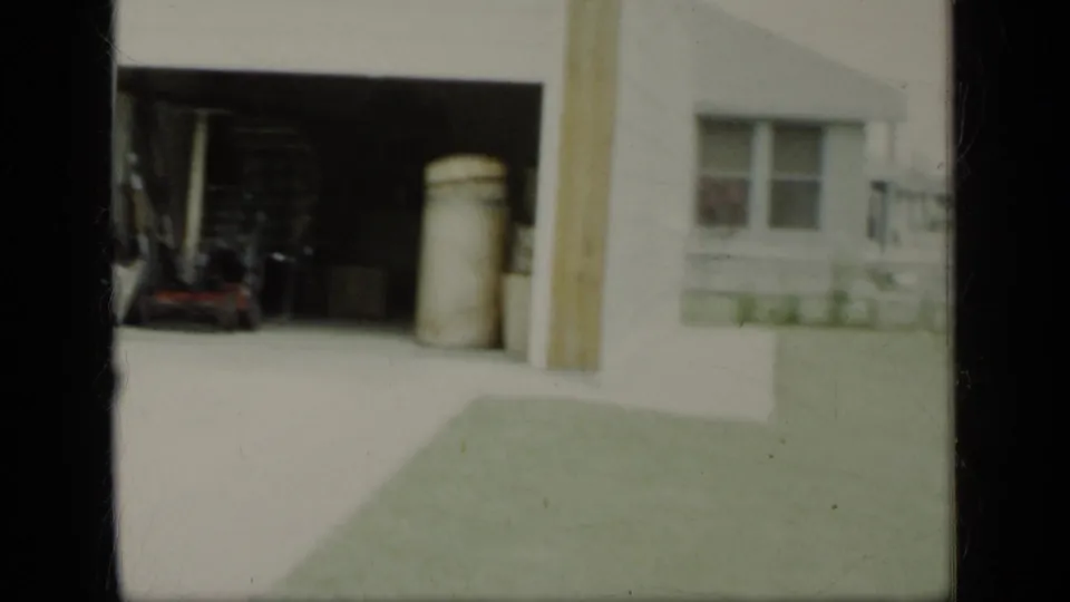 1948 New York home-movie frame showing an open garage bay, large barrel, driveway, lawn, and house exterior.