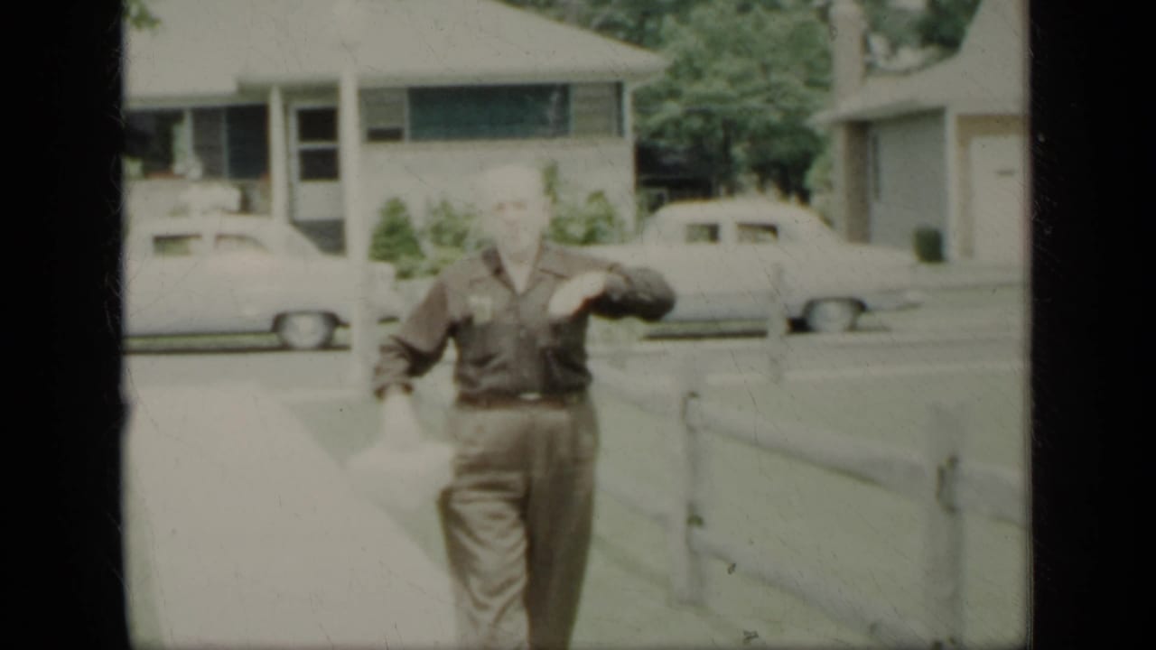 Adult man walking on a sidewalk near a fence and parked cars.