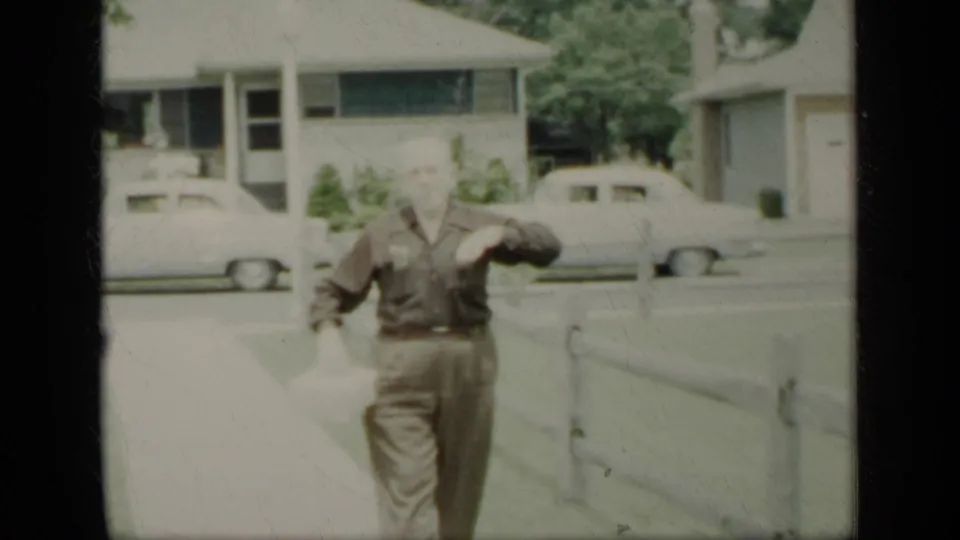 Adult man in dark shirt and trousers walks toward the camera on a New York residential sidewalk with parked cars behind him.