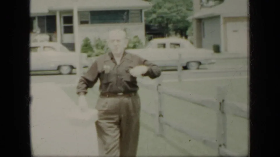 The man continues toward the camera, holding a light object and passing a low fence on a residential New York sidewalk.