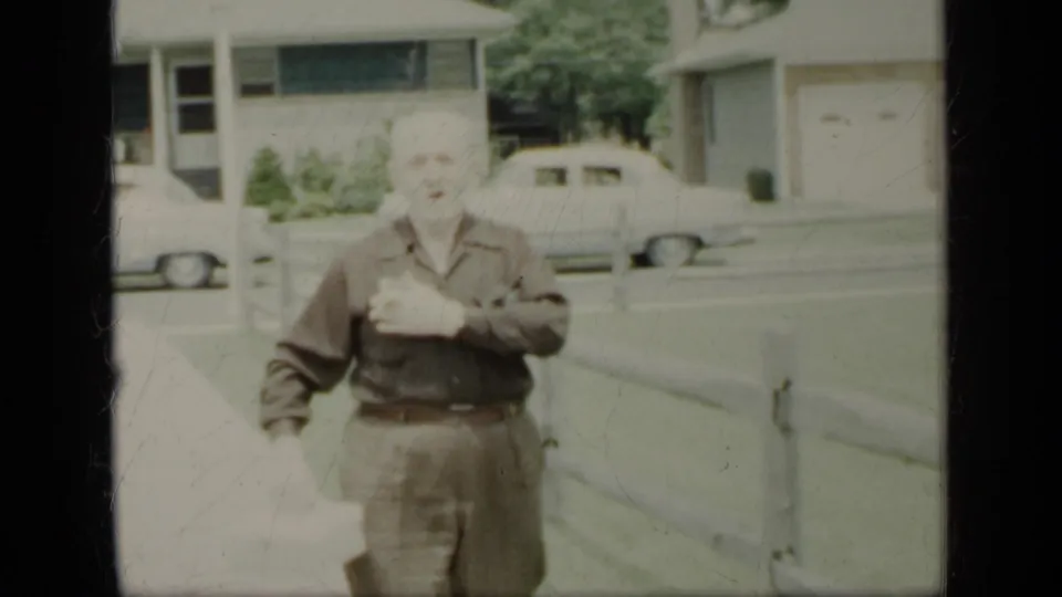 The man faces the camera and touches or adjusts his shirt, with a fence, house, and parked car in the residential background.