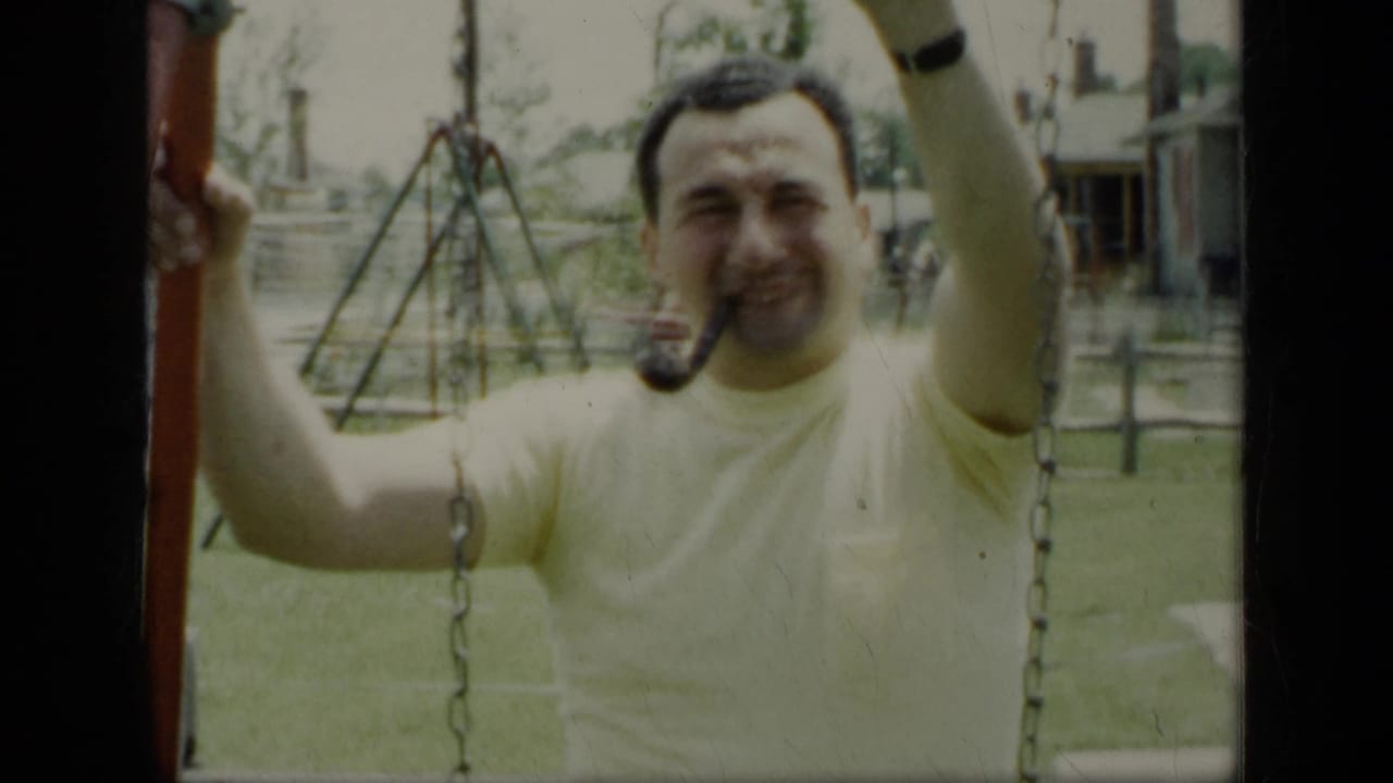 The man smiles toward the camera while gripping the swing set, with backyard structures behind him.