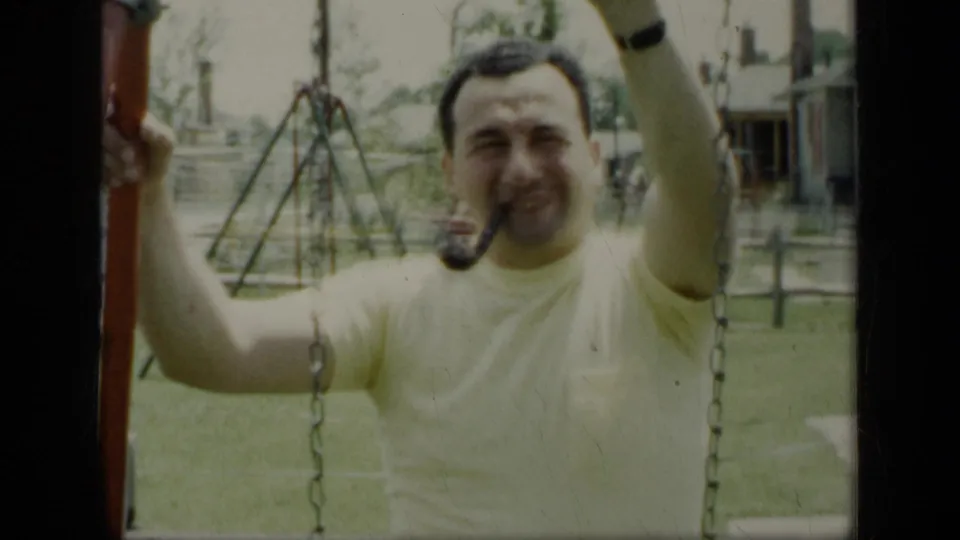 The man smiles toward the camera while gripping the swing set, with backyard structures behind him.