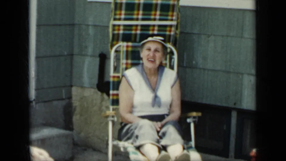 An older woman sits outdoors in a plaid folding chair beside exterior house siding in the New York home-movie footage.