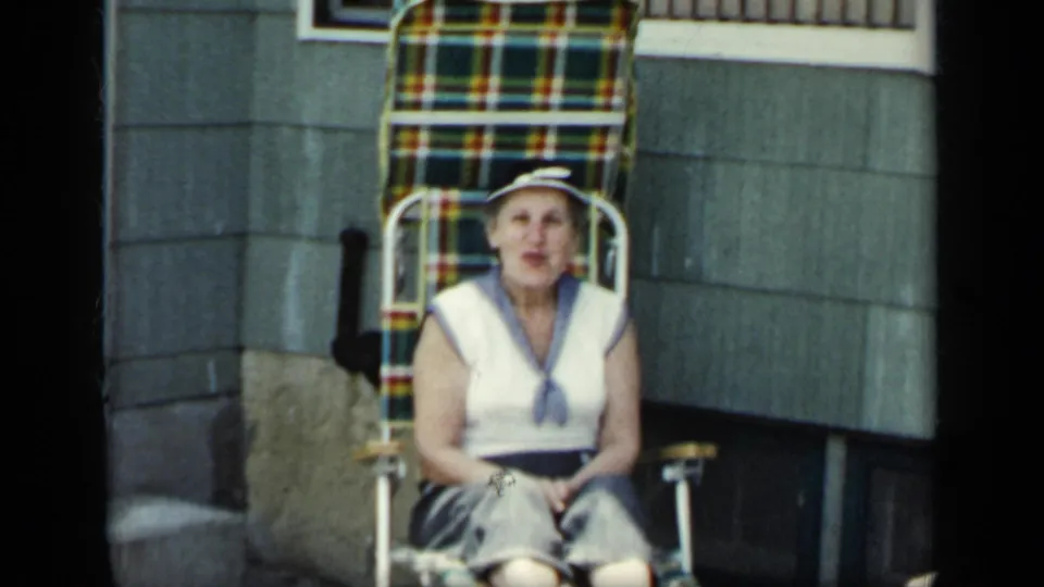 The seated older woman faces the camera from a plaid folding chair near house siding, in softly exposed 8mm color.