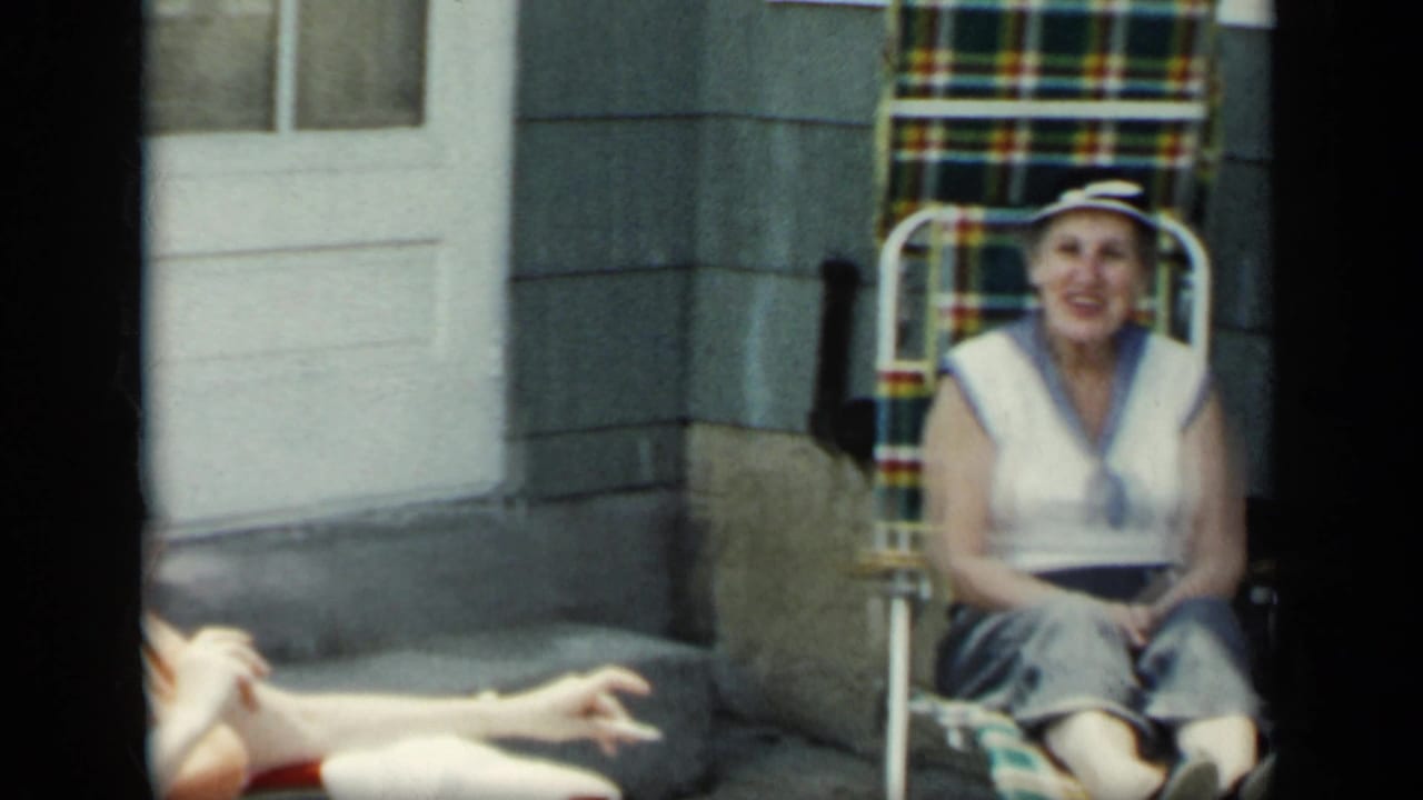 Two women sit outside a house on lawn chairs, with one centered in a white hat and sleeveless top.