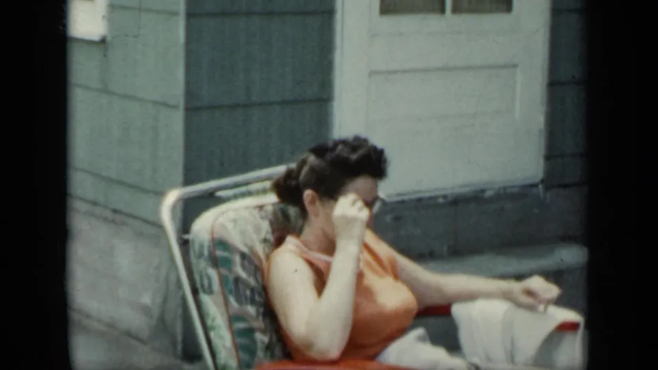 1948 New York porch frame of a seated woman in an orange sleeveless top adjusting or removing sunglasses near a white door.