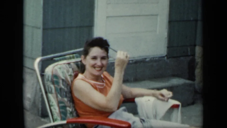 1948 New York porch frame of a smiling woman seated in a lawn chair after lifting her sunglasses toward the camera.