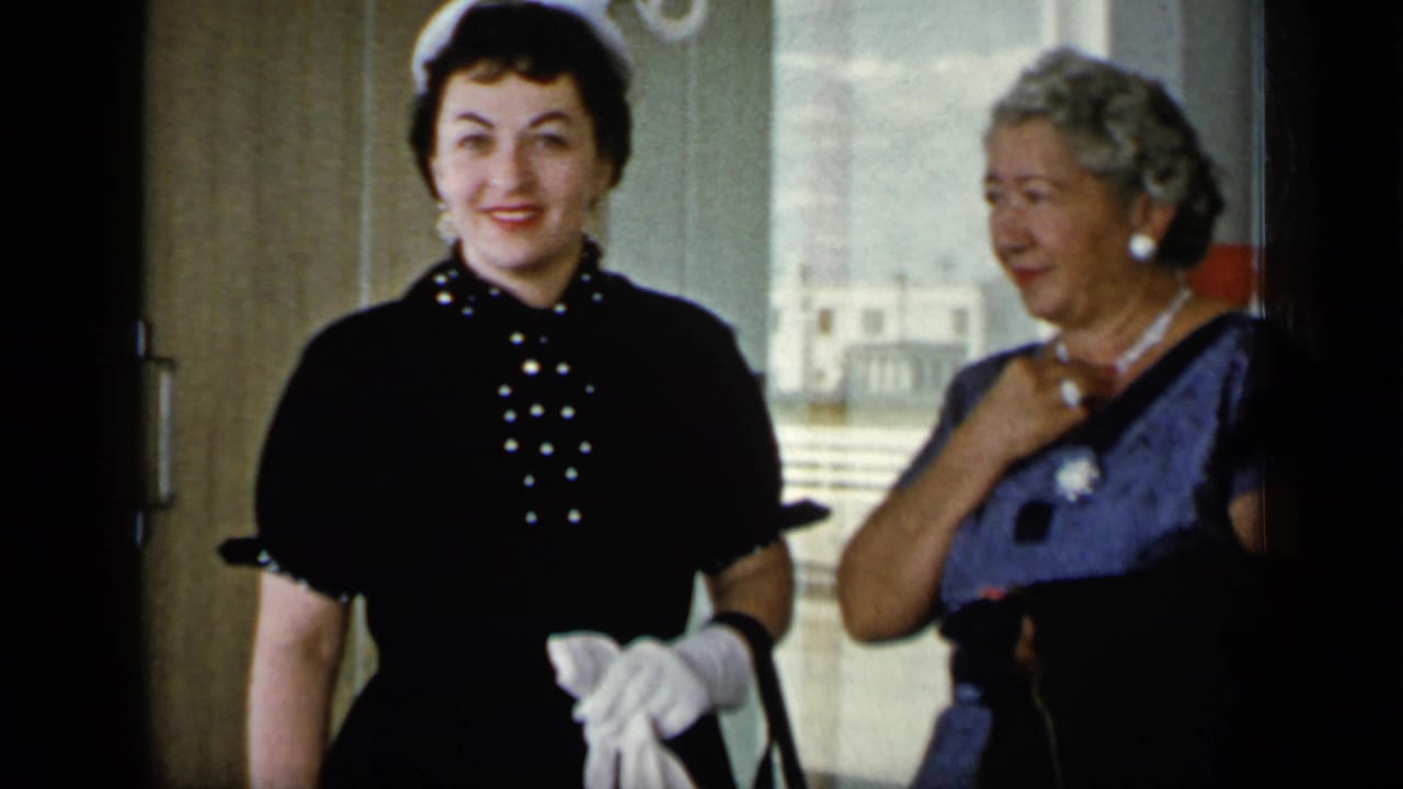 In 1948 New York home-movie footage, two women in gloves and mid-century dresses stand indoors near a bright doorway.