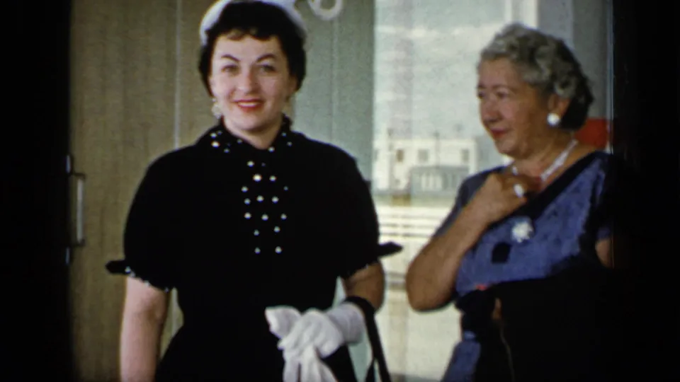 In 1948 New York home-movie footage, two women in gloves and mid-century dresses stand indoors near a bright doorway.