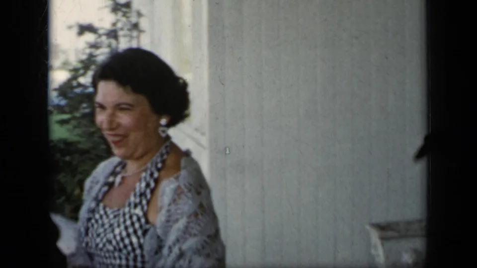 A woman wearing a checkered dress, earrings, and a light lace cardigan smiles near a window in the 1948 home-movie scene.