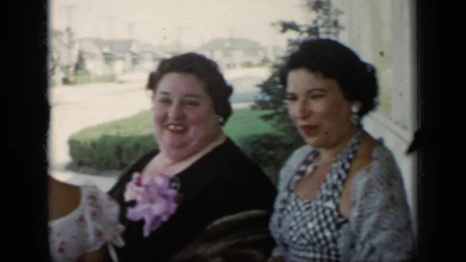 Two women in dressy mid-century clothing appear near a window, with a street and houses visible outside in the 1948 New York footage.