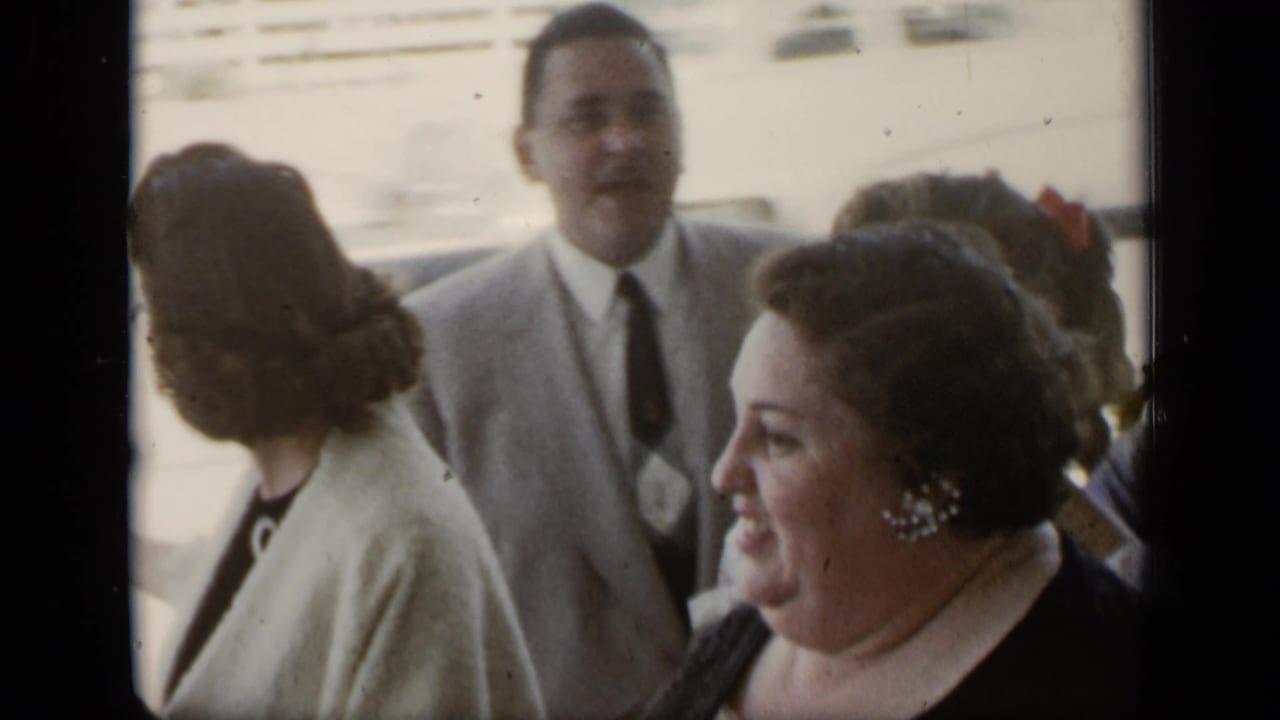Well-dressed adults stand together outdoors, including a man in a light suit and women in dark dresses and light wraps.
