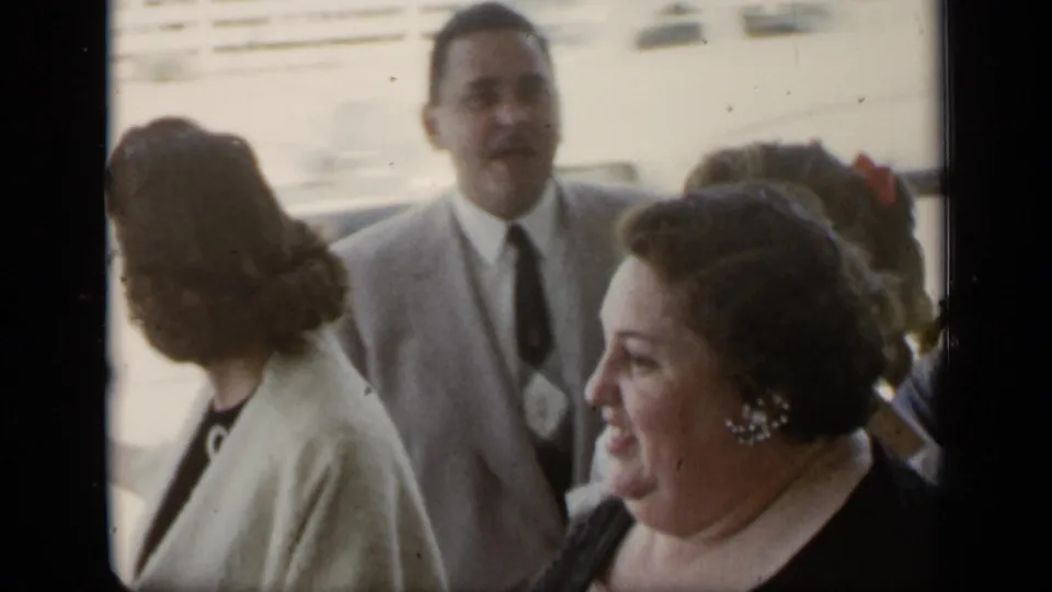 1948 New York home-movie frame of adults at an outdoor reception, with a man in a light suit and women in mid-century dress conversing nearby.