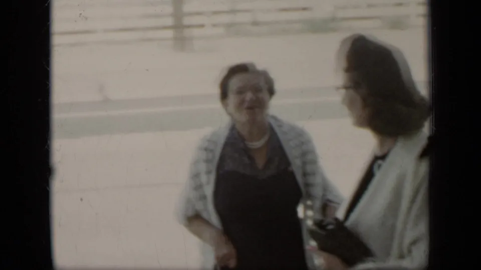 Two women at a 1948 New York outdoor gathering converse near a bright walkway, with one holding a small purse and wearing glasses.