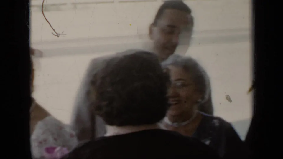 Adults at a 1948 New York reception stand close together in candid home-movie footage, with smiling faces and a man in a light suit behind them.