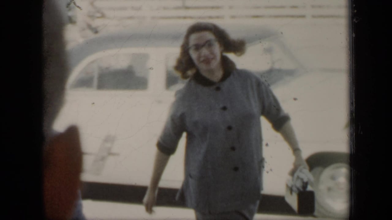 Woman in glasses and a gray button dress smiles while walking outdoors near a parked car.