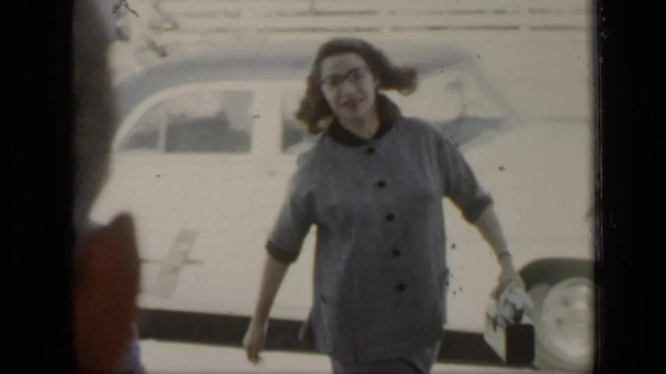 In 1948 New York home-movie footage, a woman in glasses and a gray button dress smiles toward the camera near a parked car.