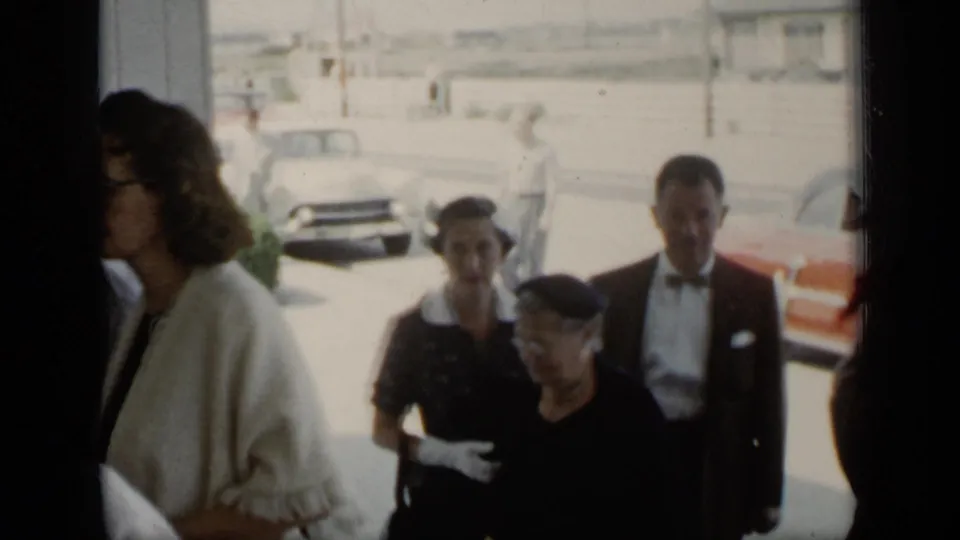 A small group of adults in 1940s dress, including a woman in gloves and a man in formal wear, walks near parked cars in New York.