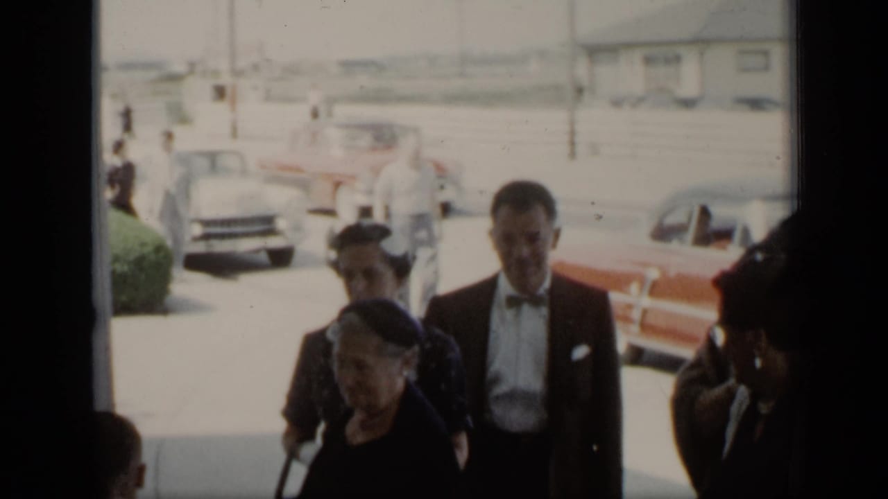 Adults in formal clothing stand near parked cars on a bright street.