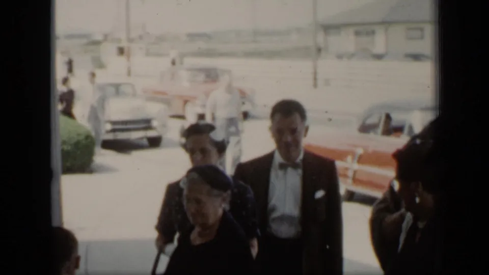In 1948 New York home-movie footage, well-dressed adults gather near parked cars on a sunlit street.