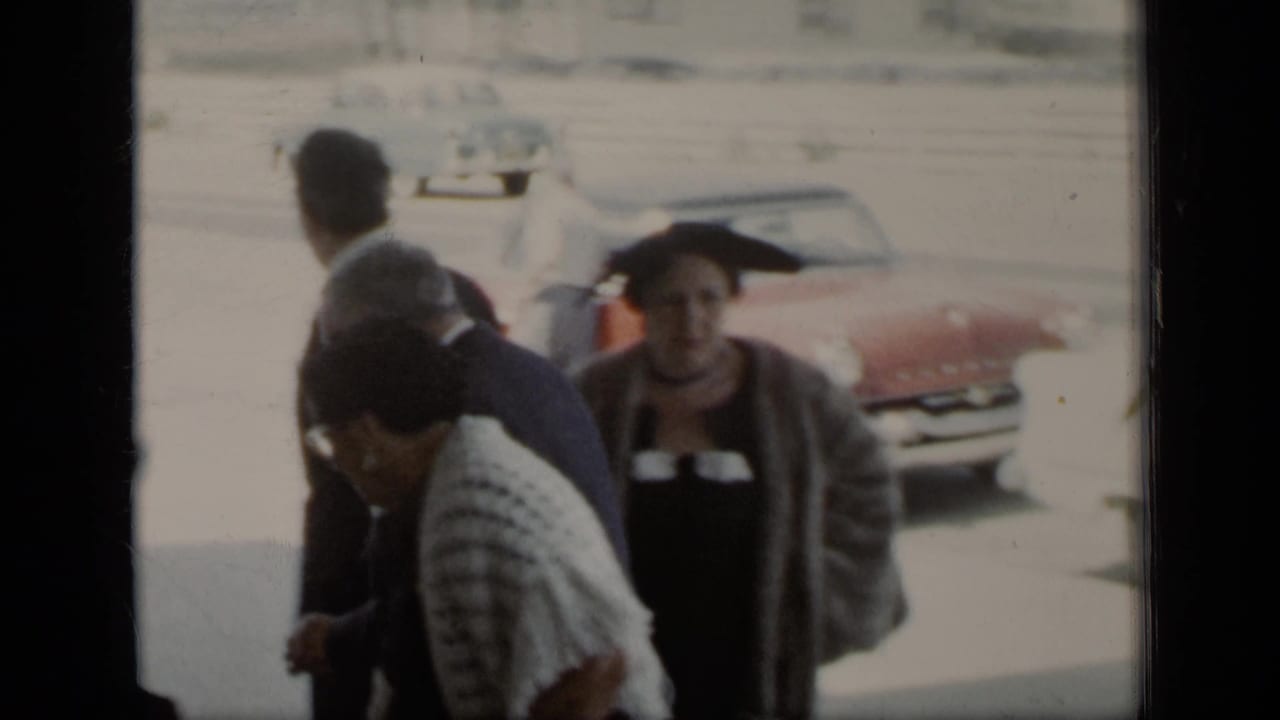 Adults in formal clothing walk near parked mid-century cars.