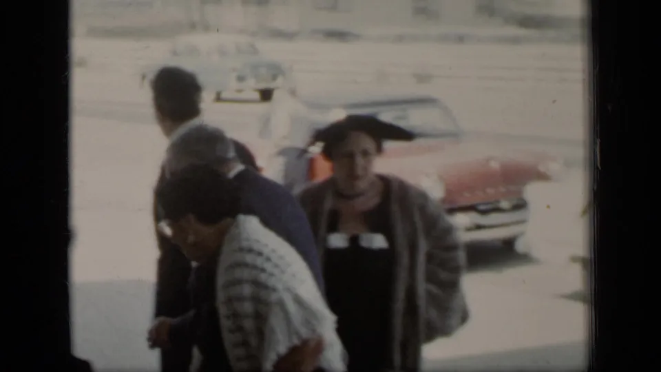 In supported 1948 New York home-movie footage, adults in suits, dresses, and hats gather outdoors near parked cars.