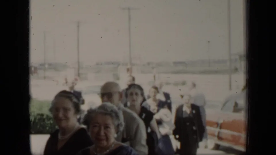 1948 New York home-movie still showing two women in the foreground with adults behind them near mid-century automobiles.