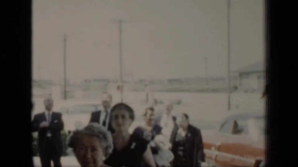 1948 New York frame showing adults moving through a bright roadside setting with classic cars and utility poles behind them.