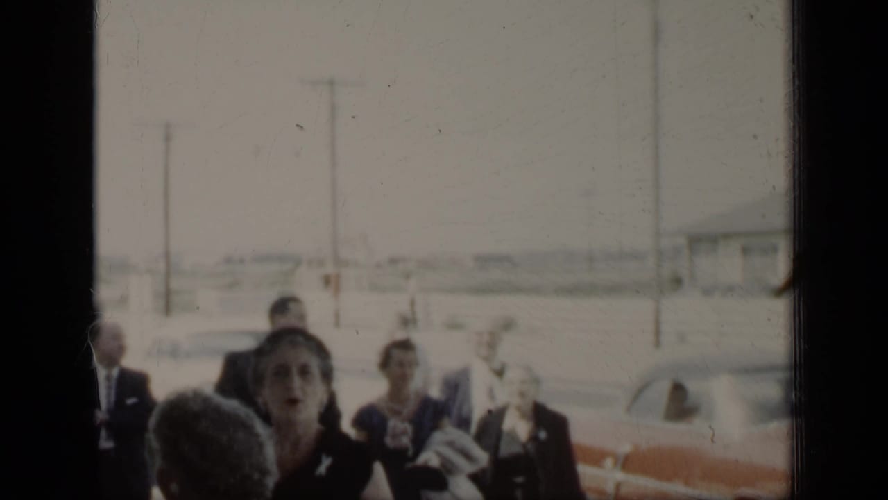Adults in formal clothing stand near parked cars and utility poles in a soft 8mm frame.