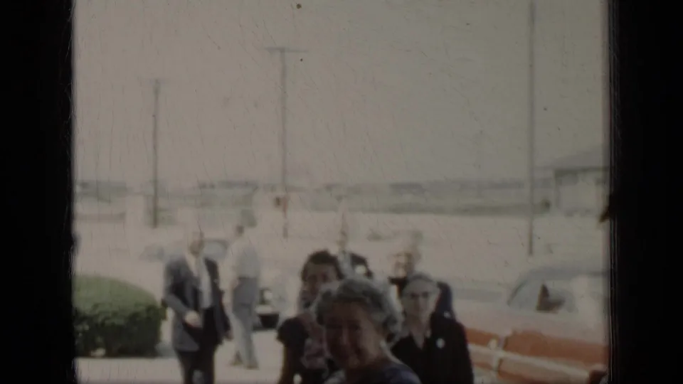 Formally dressed adults appear near hedges and cars in a grainy New York 1948 home-movie frame.